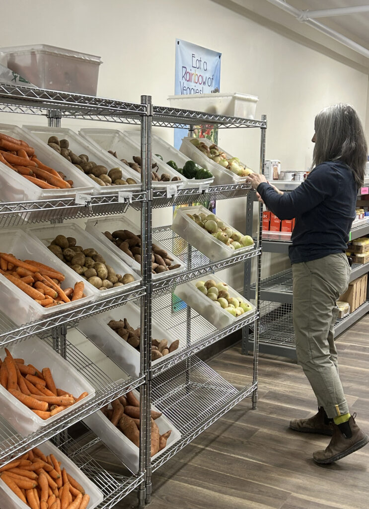 A woman checks plastic crates of produce, displayed on a wire shelving rack.