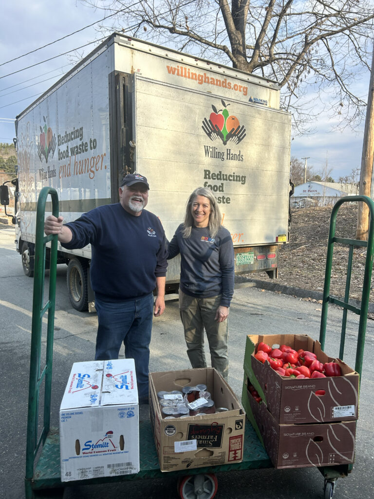 An older man with a white beard and a woman with shoulder length silver hair pose behind a cart loaded with cardboard produce boxes. A Willing Hands box truck is visible in the background.