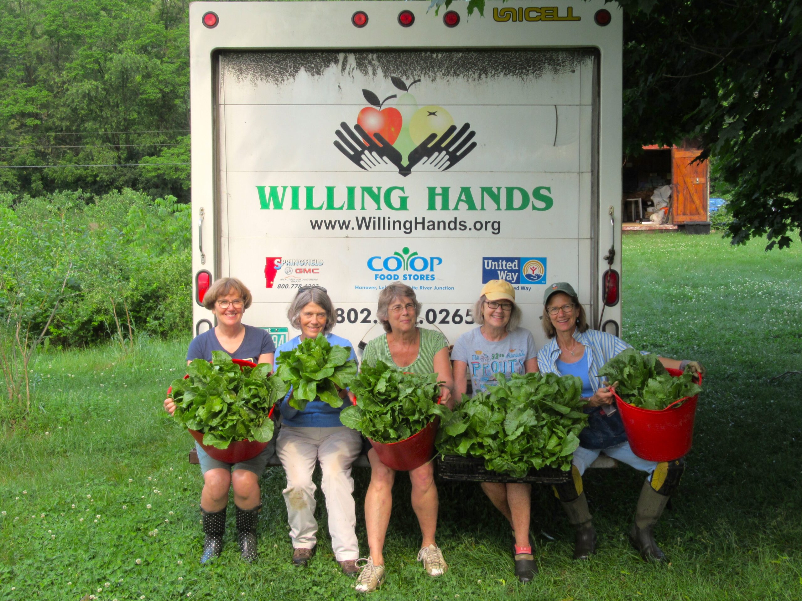 Five women sit on the back of a Willing Hands white box truck parked in a field. They are holding large bunches of fresh, green lettuce.