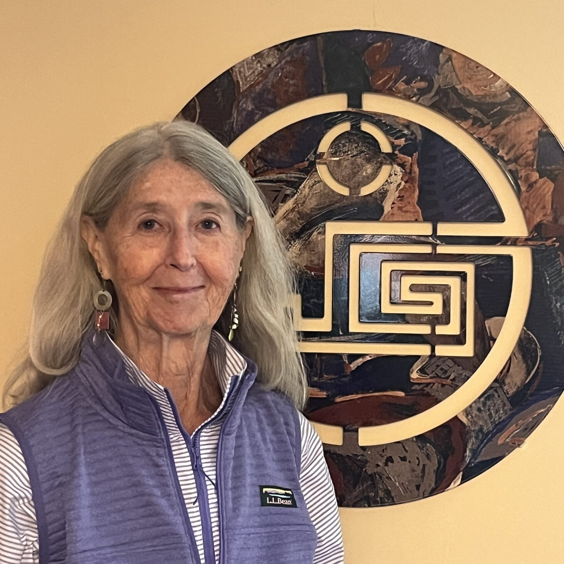 A white women with long, gray hair is standing in front of a decoration hung on a wall. She is wearing a long-sleeve shirt underneath a purple sport vest.