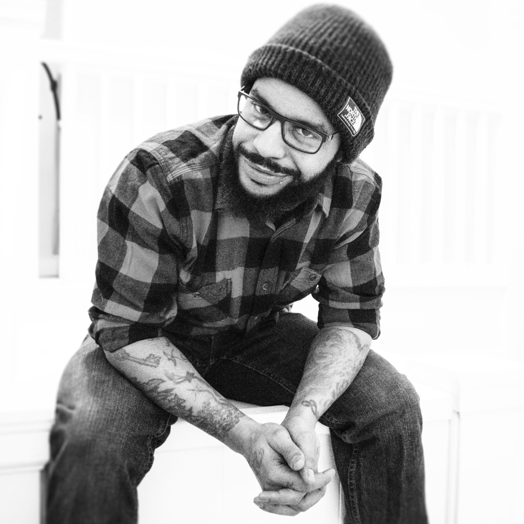 A black and white photo of a smiling man in a beanie hat, plaid shirt, and jeans. He has glasses and a mustache and beard. He is sitting with elbows on thighs, and leaning forward with clasped hands.