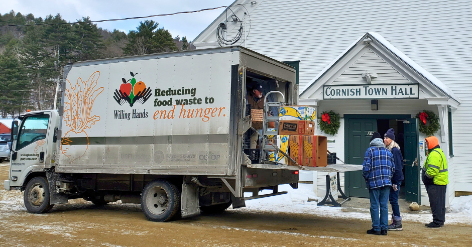 Boxes of food are unloaded from a Willing Hands delivery truck, in front of a large white building with a sign reading "Cornish Town Hall."