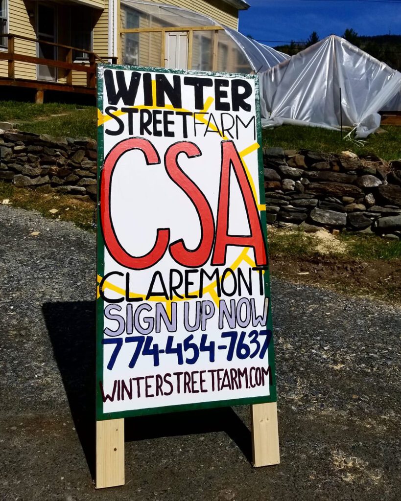 A hand painted sign displayed on a gravel driveway reading "Winter Street Farm CSA Claremont. Sign up now. 774 454 7637. winterstreetfarm.com
