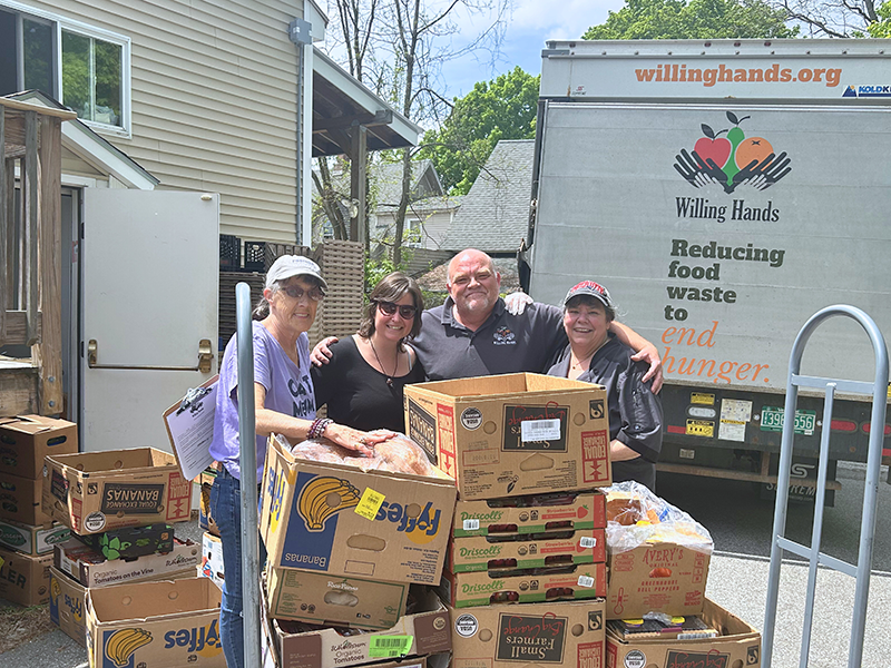 Tim, Beth, and Claremont Soup Kitchen Staff 2024 edit Four people pose behind a hand cart loaded with cardboard boxes of food. A tan building and box truck with the Willing Hands logo are in the background.