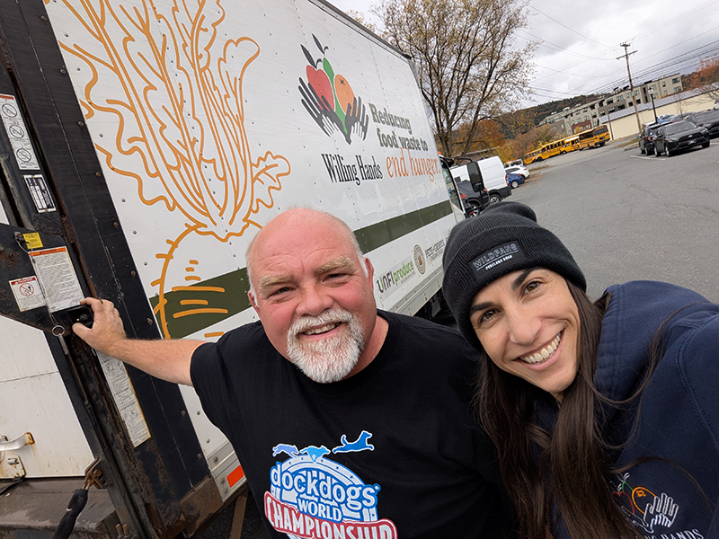 Delivery Coordinator Tim and board member Jo Mirenda A man with a white beard wearing a black teeshirt, and a woman with long dark hair wearing a beanie and hoodie, pose for a selfie in front of a box truck with the Willing Hands logo on it