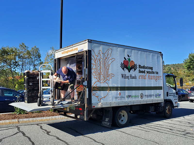 A man stands in the back of a box truck marked with the willing hands logo, weighing a cardboard box on a large scale.