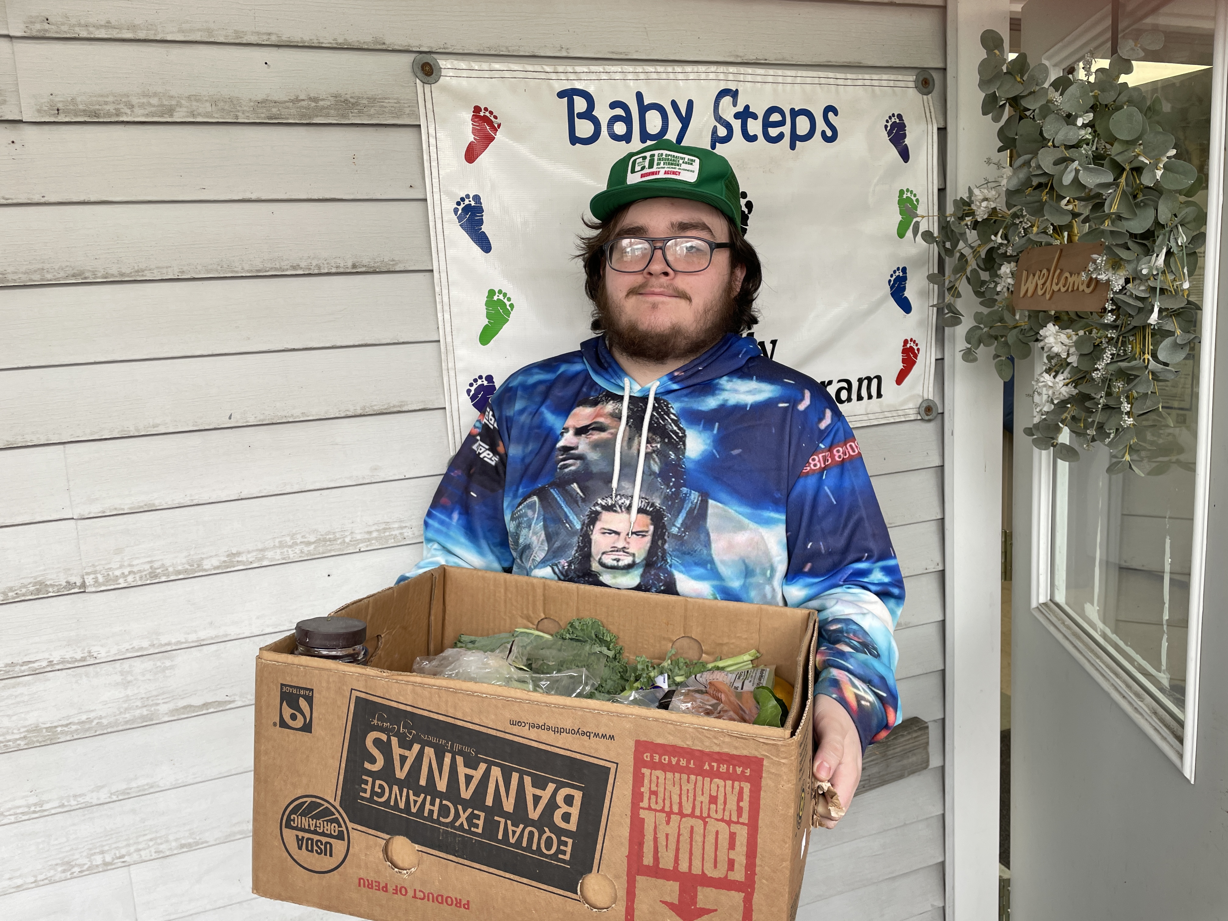 A young man in a colorful hoodie and ball cap holds a cardboard box of produce in front of a sign reading "baby steps"
