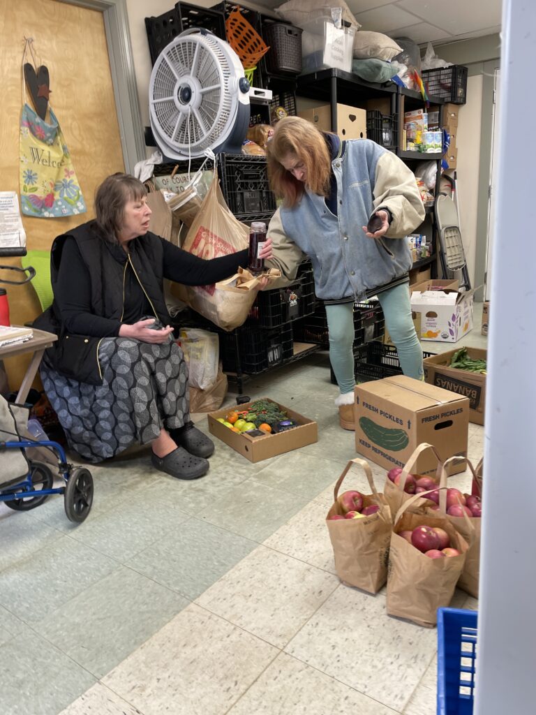 A seated woman and a standing woman pass a class jar between each other in a room crowded with boxes of produce and other food.