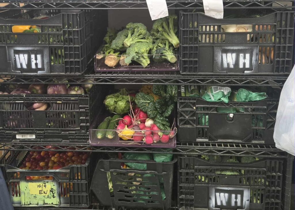 A stack of produce crates, many labeled with WH, with loaves of bread and bags of greens stacked on top.