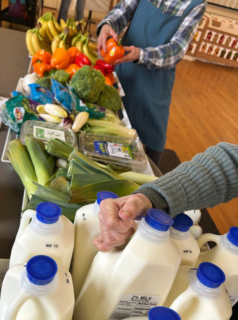 A hand sets a milk jug down on a table. In the background, another figure arranges produce on the table
