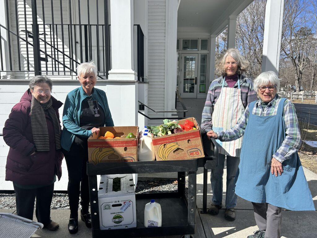 Four women in coats and aprons pose on either side of a cart loaded with boxes of fresh produce