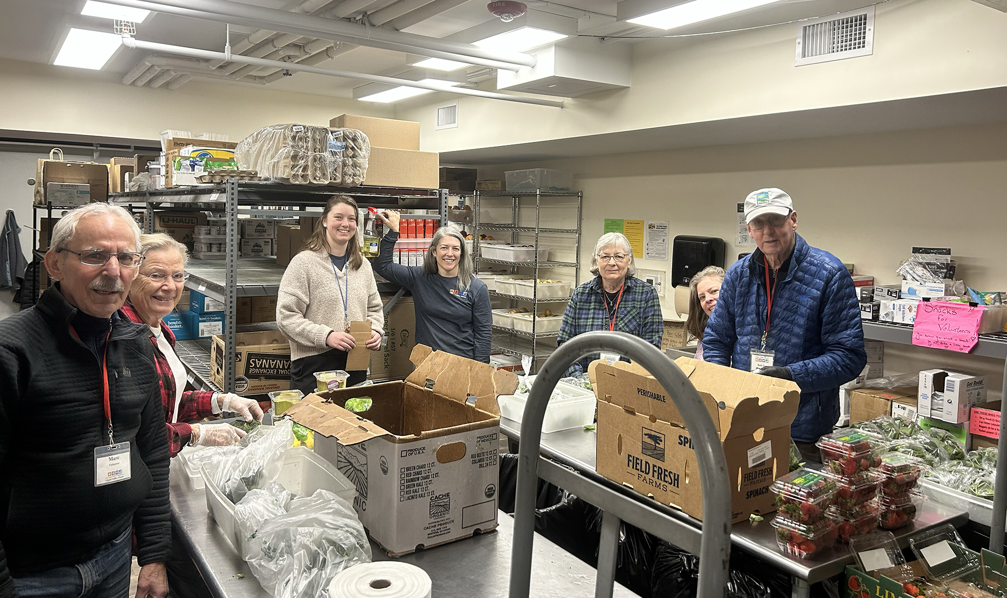 Seven adults pose around metal tables loaded with boxes of produce. The background shows shelves of food in a room with fluorescent lights