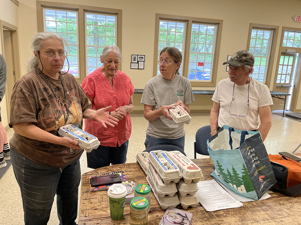 West Fairlee Food Shelf - Volunteers with eggs Three older women and one man stand around a table with several cartons of eggs on it. Two women hold cartons of eggs. One woman appeals to be speaking to the photographer. The others look at her.