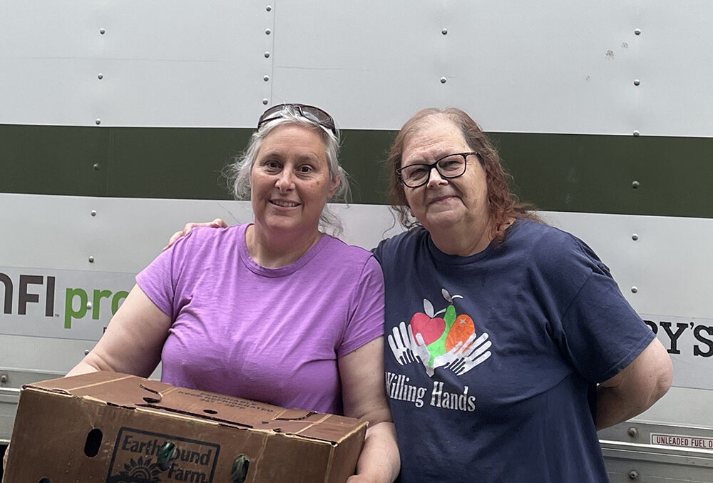 West Fairlee Food Shelf - Jen Shatney and Pamela Mainville Two women pose together for a photo. The one one oneleft has grey hair pulled back, is wearing a purple teeshirt, and is holding a cardboard produce box. The one one the right wears glasses, is wearing a navy blue "Willing Hands" teeshirt, and has her arm around the other woman.