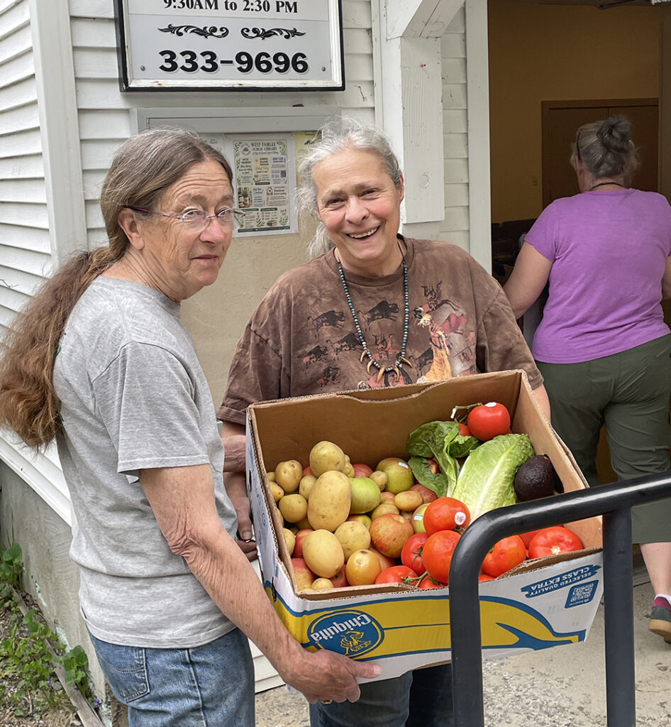 West Fairlee Food Shelf - Laurie and Lynn Two women pose with a banana box full of mixed produce in front of a white clapboard building with a sign reading "West Fairlee Town Offices"