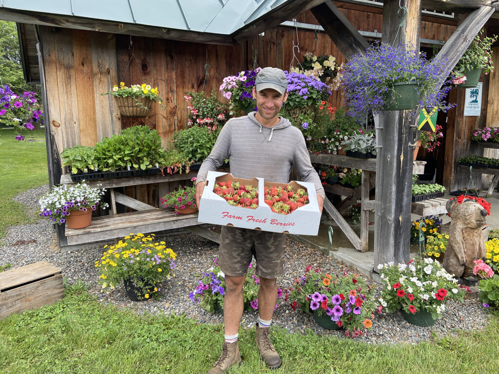 Phil-Mason_Crossroad_IMG_8396 A smiling man in a grey shirt, ball cap, shorts, and boots, holds a flat of strawberries in from of a display of hanging flower baskets