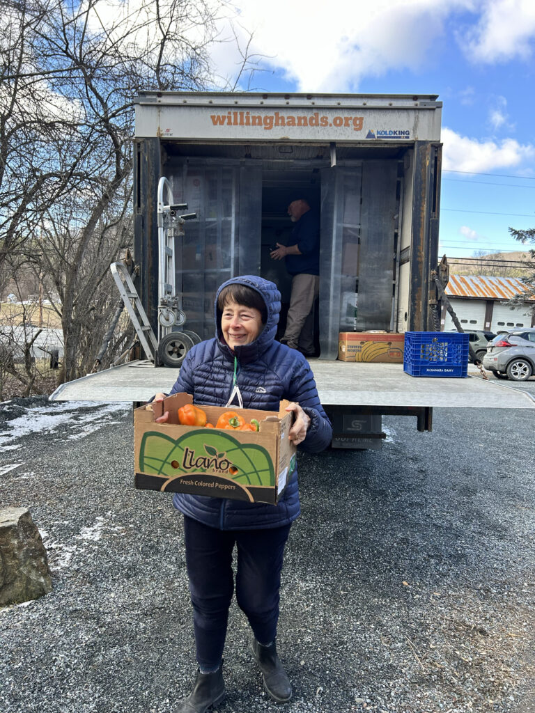 A smiling woman in a winter coat carries a box of orange peppers away from the back of the Willing Hands delivery truck
