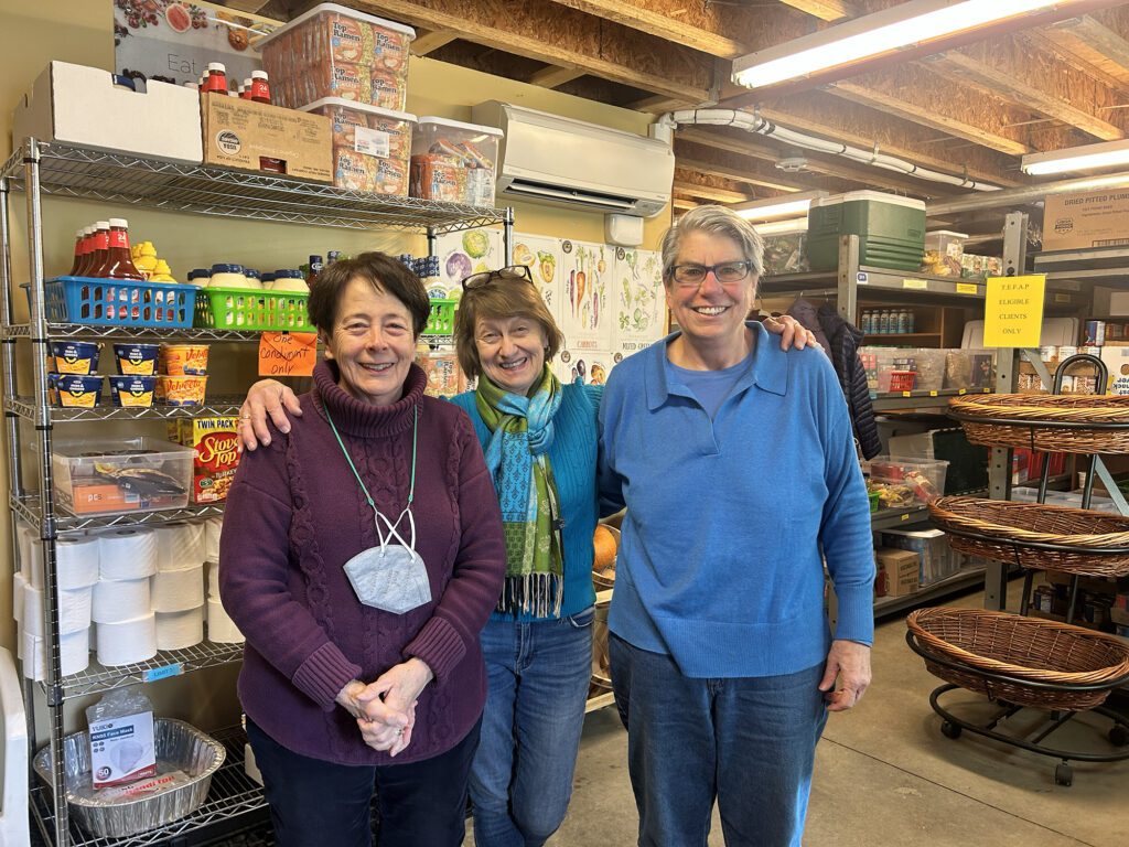 three smiling women stand together in front of wire shelves stacked with food