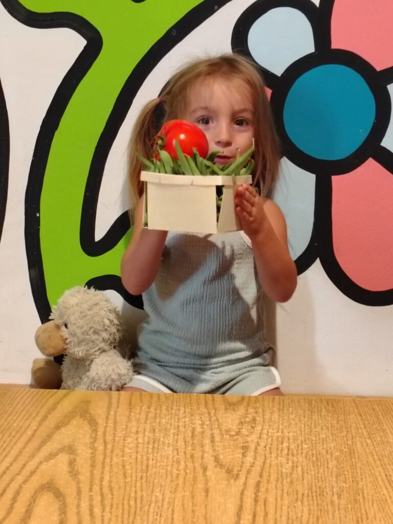 A girl with blond pigtails holds a carton og greenbeans, topped with a large red tomato, up in front of her face.