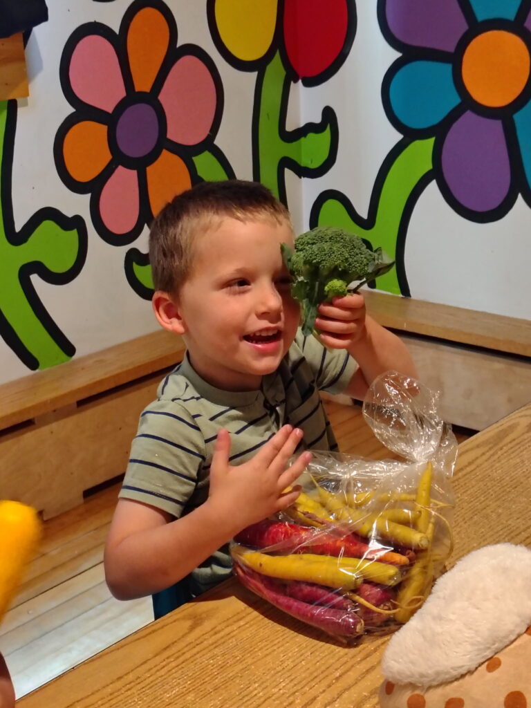 A little boy holds a broccoli crown in front of his face, while a bag of colorful carrots sits in front of him on a table.  