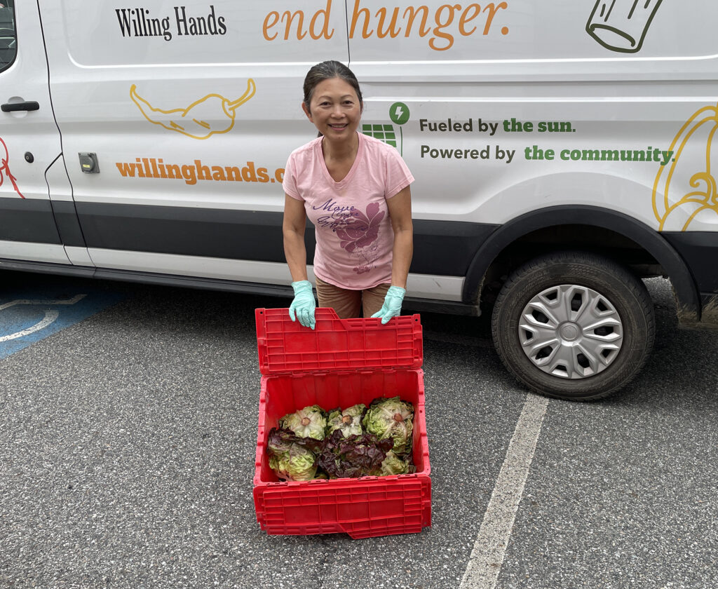 A woman in a pink teeshirt poses in front of a Willing Hands van with a red plastic crate of lettuce