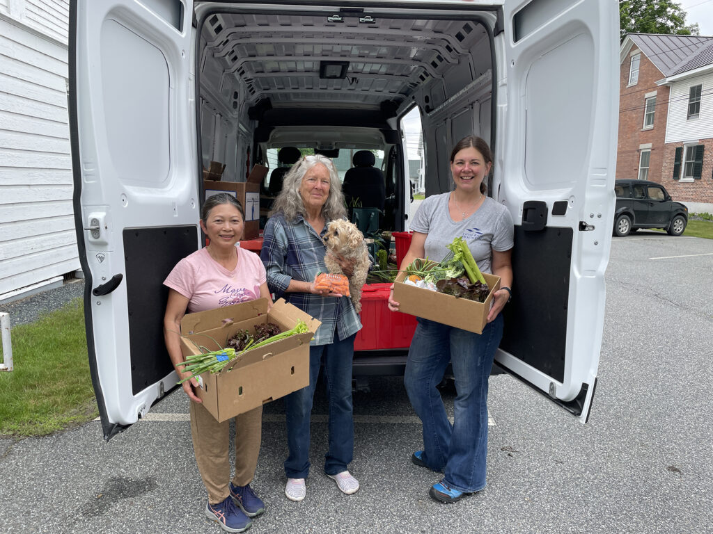 Three women pose with boxes of fresh produce between the open back doors of a delivery van.