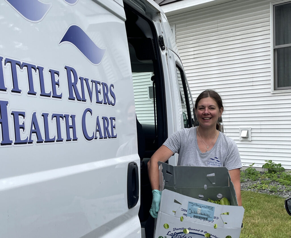 A woman stands beside a white van with "little rivers health care" written on the side. She showed off a large cardboard box of produce.