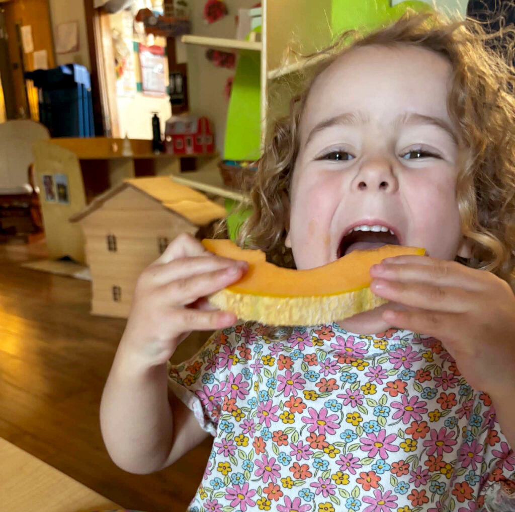 A little girl with pale skin, and curly hair takes a giant bite of melon